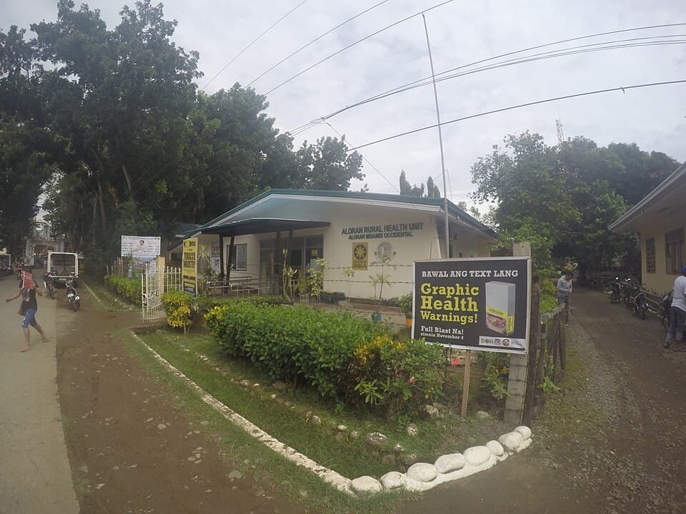 The Aloran Rural Health Unit in Aloran, Misamis Occidental, a single-storey government health building behind a hedge and a roadside health sign