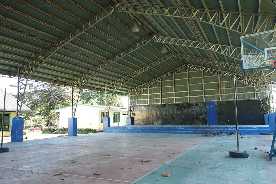 The inside of an open-sided covered court at Banga Central Elementary School in Banga, South Cotabato, with a steel truss roof, a basketball goal and a stage at the far end