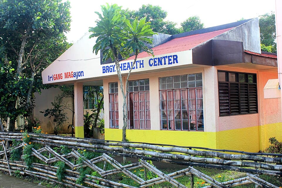 A single-story barangay health center with blue lettering, yellow and pink walls and a bamboo fence in front, located in San Ramon, Iriga City, shown as a representative example