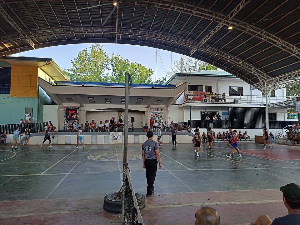Young men playing a basketball game under the roof of the Alubijid Municipal Covered Court in Misamis Oriental, with spectators seated along the stage and sidelines
