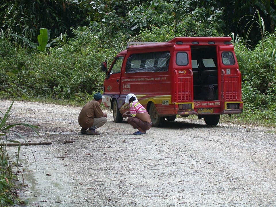 Two people crouching on a gravel road beside a parked red passenger vehicle, with thick forest behind them, photographed in the Bislig area in 2003