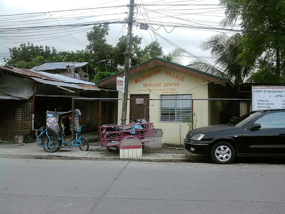 A small cream-colored barangay day care center building with a red-lettered sign beside a street in La Paz, Iloilo City, shown as a representative example of a Philippine day care center