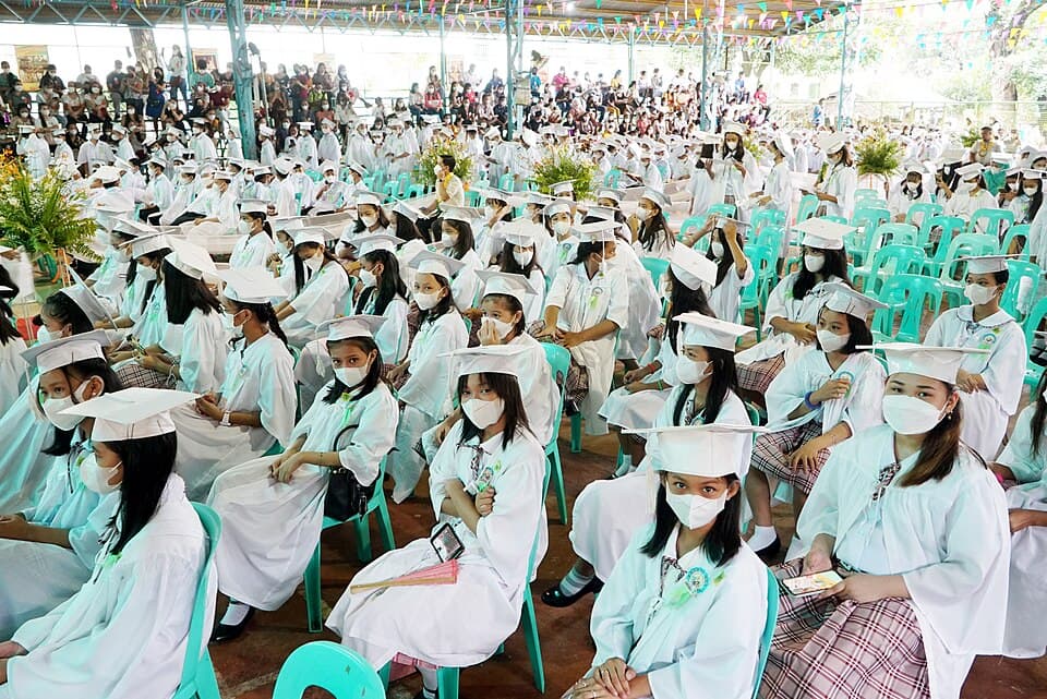 Students in white graduation togas and caps seated in rows at a school graduation ceremony in a covered court in Caloocan City, July 2022