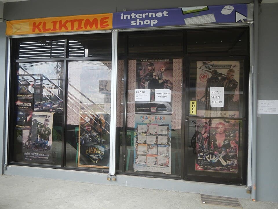 The glass storefront of a small internet shop in Bustos, Bulacan, with game posters and signs offering e-load, printing and photocopying, shown as a representative example of a Philippine internet cafe
