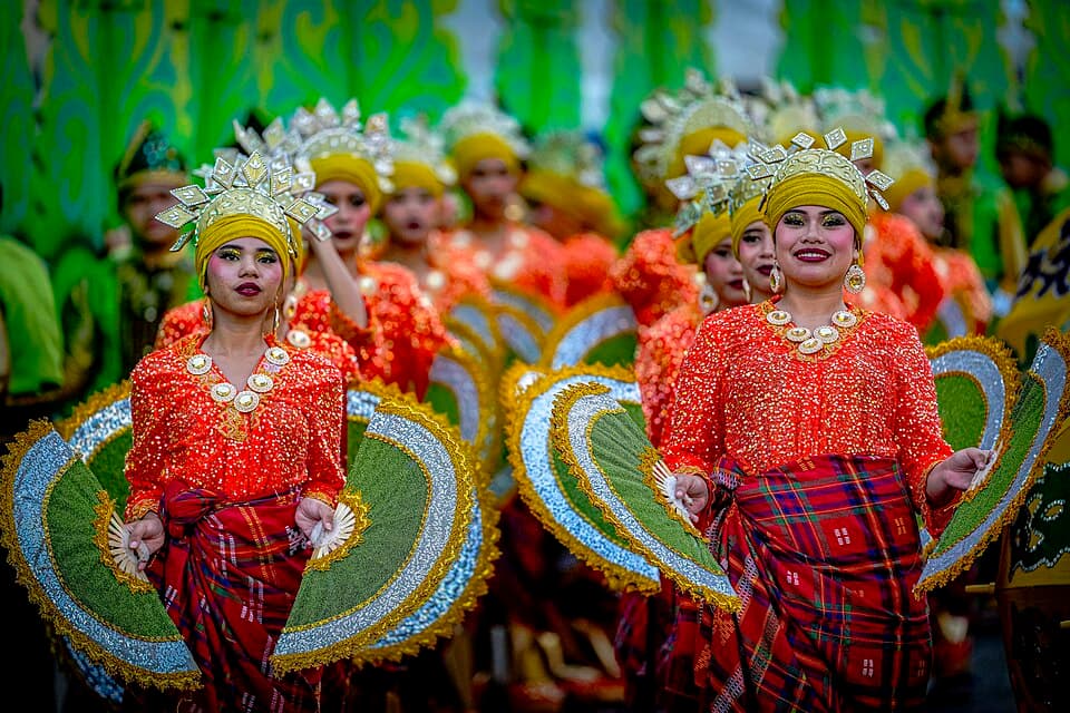 Festival dancers in red sequined blouses, plaid skirts and gold headdresses holding green fan props during the 2026 Kadayawan Festival street parade in Davao City