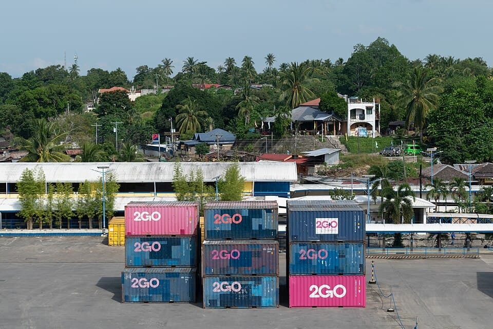 Stacks of pink and blue 2GO shipping containers on the concrete yard of the Port of Nasipit in Agusan del Norte, Mindanao, with port sheds and hillside houses behind