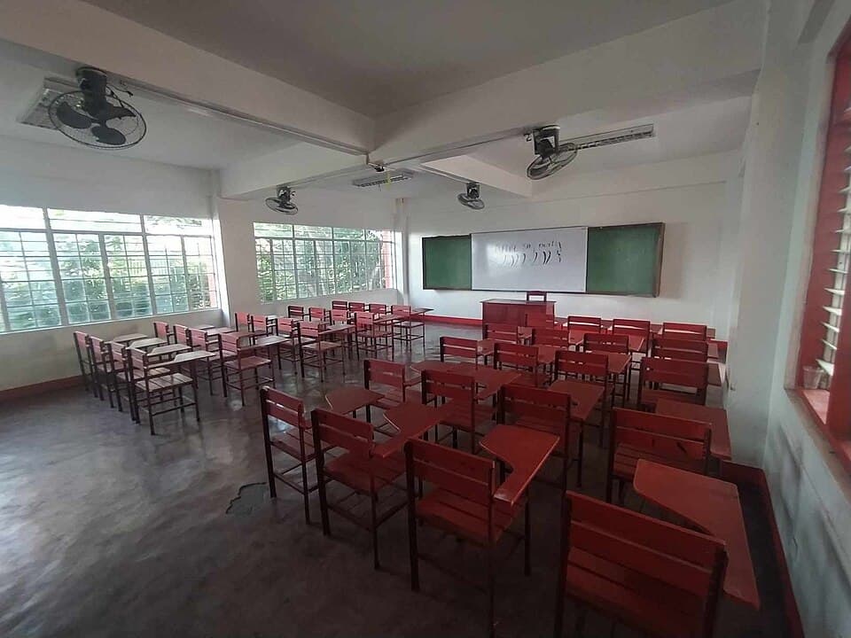An empty public high school classroom with rows of red wooden armchairs, ceiling fans and a whiteboard between two green chalkboards, at Western Bicutan National High School in Taguig