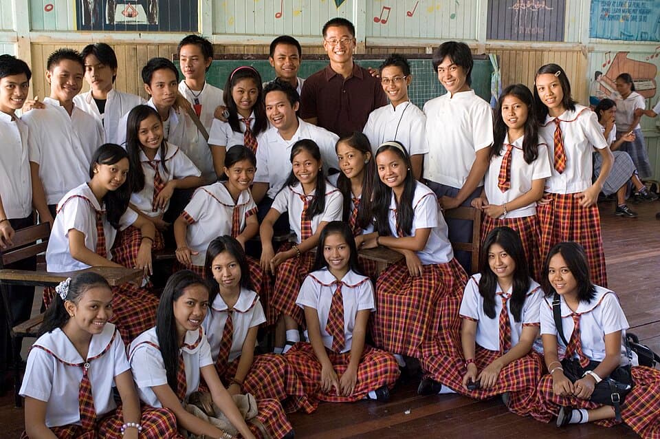 A teacher poses with a class of Filipino high school students in their school uniforms inside a wooden classroom in the Philippines