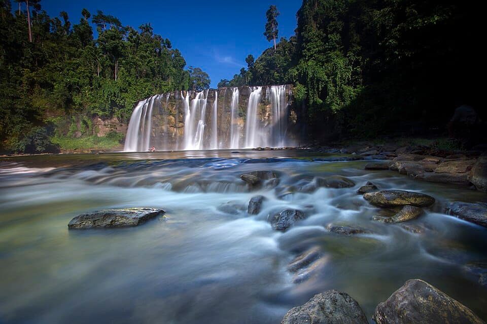 Long-exposure photo of Tinuy-an Falls in Barangay Borboanan, Bislig City, with water streaming over the wide rock face and smooth rapids flowing past boulders in the foreground