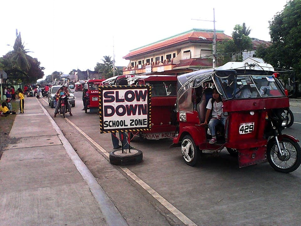 Red passenger tricycles lined up beside a Slow Down School Zone sign outside an elementary school in Sto. Tomas, not in Bislig