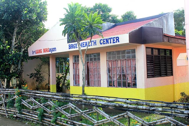 A single-story barangay health center with blue lettering, yellow and pink walls and a bamboo fence in front, located in San Ramon, Iriga City, shown as a representative example