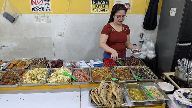 A woman serves food from trays of cooked dishes at the counter of a carinderia, a small Filipino eatery, in Cebu City