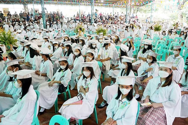 Students in white graduation togas and caps seated in rows at a school graduation ceremony in a covered court in Caloocan City, July 2022