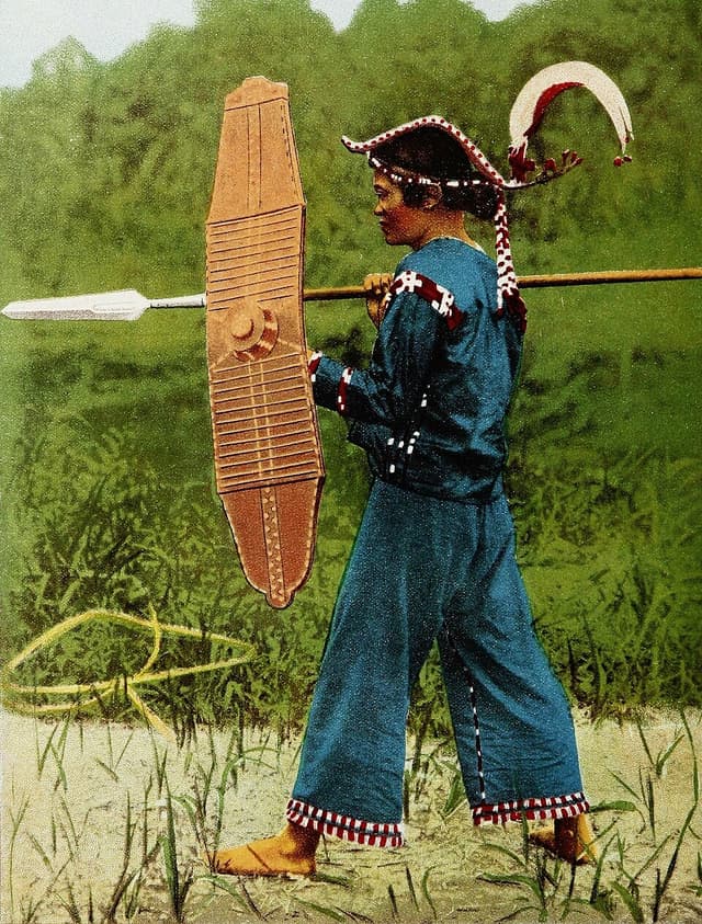 A hand-colored 1913 photograph by Dean Worcester of a Mandaya man from the Davao Gulf area, in an embroidered blue outfit and headdress, carrying a long wooden shield and a spear