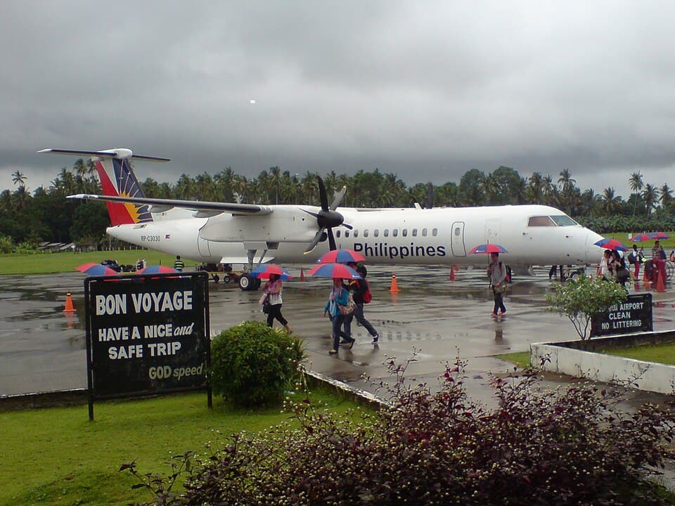 A Philippine Airlines Bombardier Dash 8 Q400 turboprop parked on a wet apron at Ozamiz Airport in Misamis Occidental as passengers with umbrellas walk to the terminal