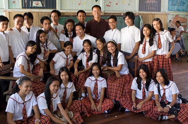 A teacher poses with a class of Filipino high school students in their school uniforms inside a wooden classroom in the Philippines