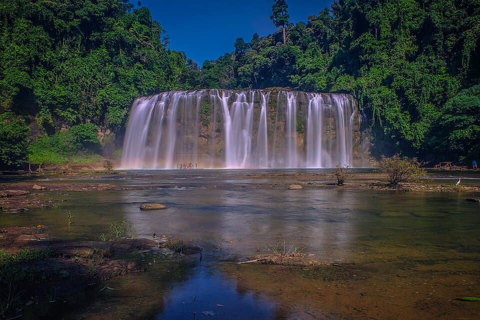 Tinuy-an Falls in Bislig City, a wide curtain of white water falling over a rock ledge into a shallow river, surrounded by dense green forest under a deep blue sky