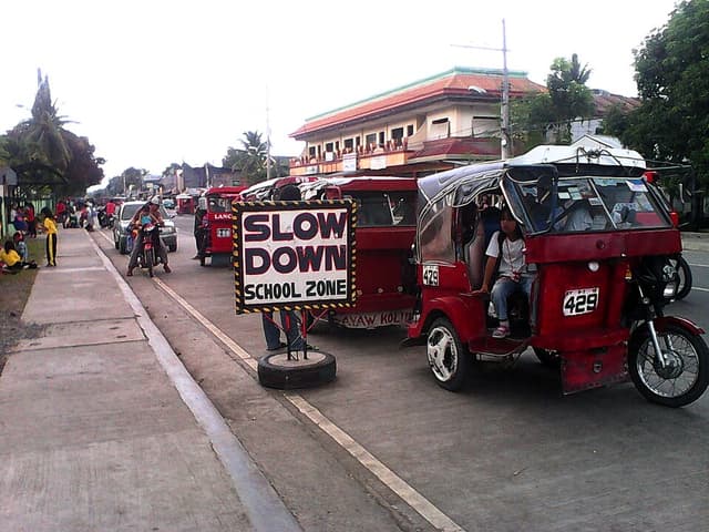Red passenger tricycles lined up beside a Slow Down School Zone sign outside an elementary school in Sto. Tomas, not in Bislig