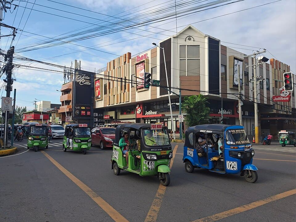 Green and blue numbered tricycles driving through a street intersection in Tagbilaran City, Bohol, in front of a commercial building with fast-food signs